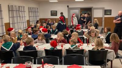 spejdere der venter på julemad, sidende ved borde med hvide duge og nissehuer på.